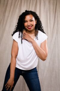 Photo of Tracey Baptiste. Black woman with long curly hair wearing a white t-shirt and jeans, smiles at the camera.